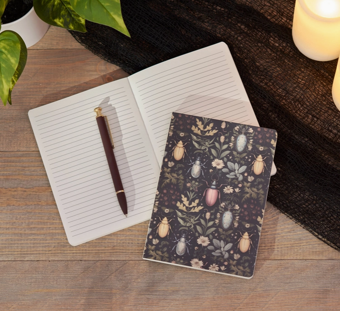 A styled flat-lay of a small notebook with a dark floral and insect pattern, placed next to an open white lined notebook with a brown pen on a wooden surface.