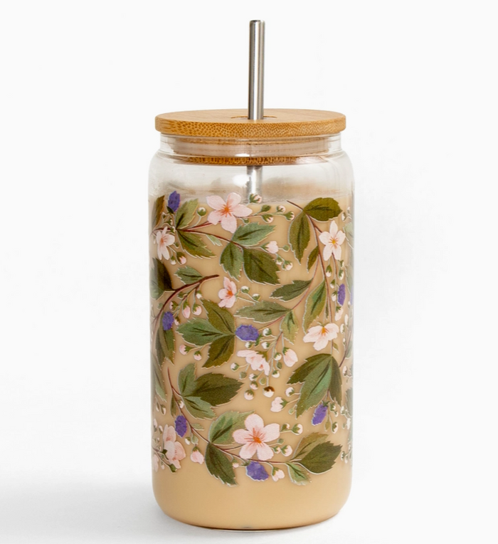 A clear glass tumbler with a bamboo lid and metal straw, decorated with a delicate floral pattern of green leaves, pink blossoms, and small purple berries, filled with a light beige beverage.