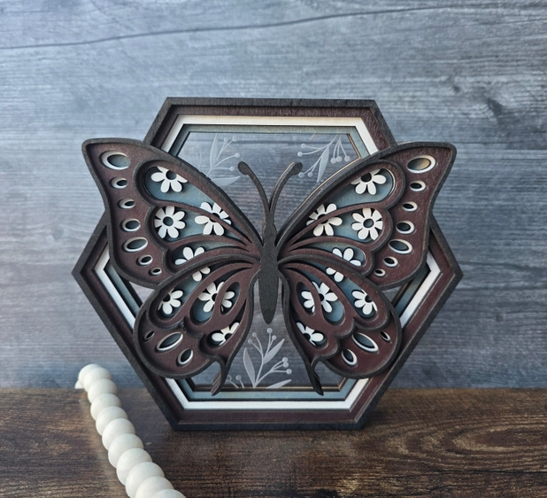 A decorative butterfly-shaped shelf sitter decor piece with layered dark brown wood and white floral inlay, displayed against a gray wood-textured background.