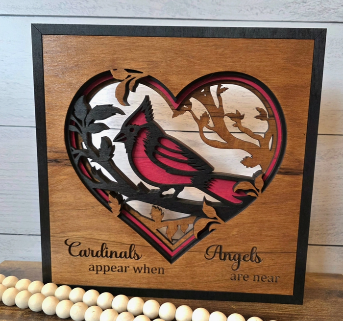 A square wooden decorative sign with a heart-shaped laser-cut design featuring a cardinal bird and the phrase 'Cardinals appear when Angels are near,' displayed on a wooden surface with a beaded garland in front.