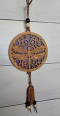 A circular wooden dragonfly wall hanging ornament with a carved dragonfly design, hanging from a brown cord with beads and a dangling leather tassel against a white wood plank background.