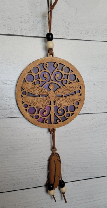 A circular wooden dragonfly wall hanging ornament with a carved dragonfly design, hanging from a brown cord with beads and a dangling leather tassel against a white wood plank background.