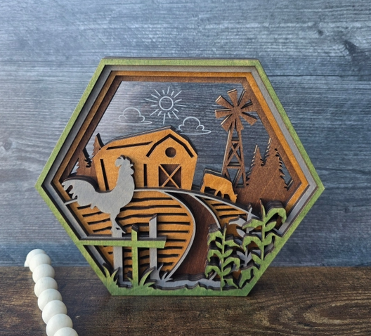 Hexagonal wooden cutout shelf sitter featuring a farm scene with a barn, windmill, trees, fence, and rooster, displayed on a rustic wood surface against a blue-gray wood plank background.