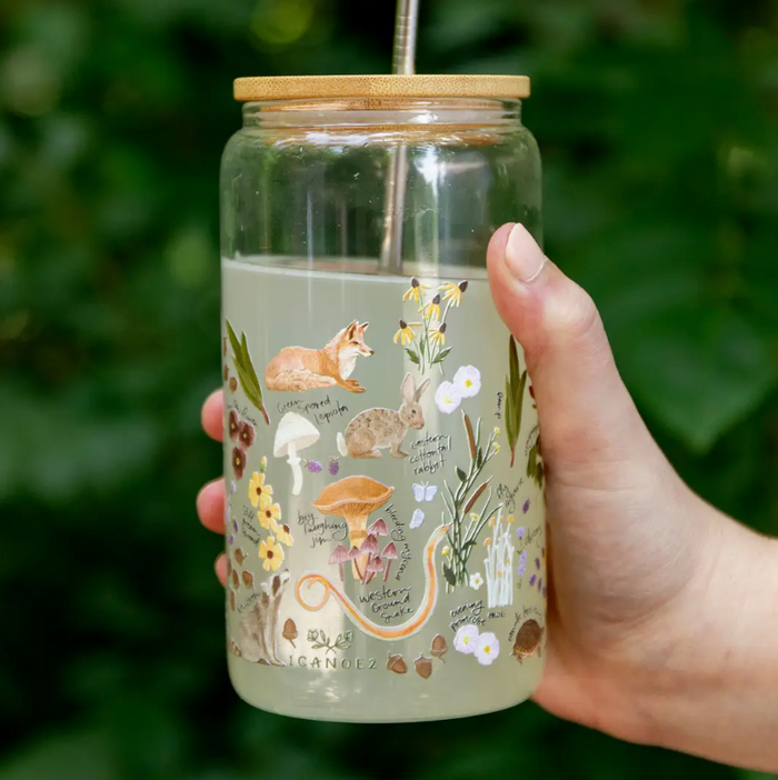 A hand holding a clear glass water bottle outdoors, featuring whimsical woodland illustrations of a fox, mushrooms, flowers, and a snake on a pale green background, with a bamboo lid and metal straw.
