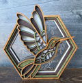 A layered wooden decorative plaque featuring a cutout hummingbird in front of a hexagonal floral frame, photographed on a rustic gray wood surface.