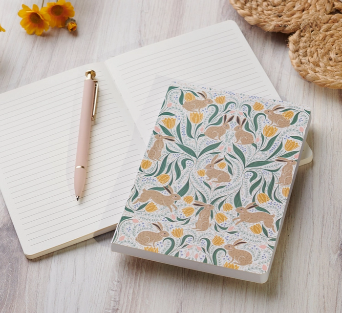 A decorative notebook with a white cover featuring a whimsical pattern of rabbits, leaves, and yellow flowers, styled on a light wood surface next to an open lined notebook, a pink pen, and small yellow flowers.