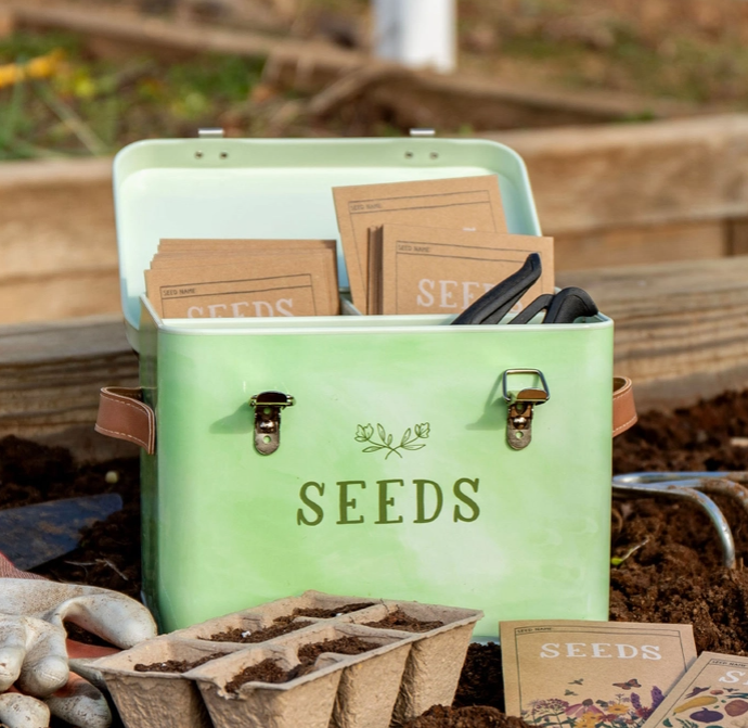 A mint green metal seed storage cooler filled with seed packets, placed outdoors in a garden bed with soil, gardening gloves, and seed trays visible around it.